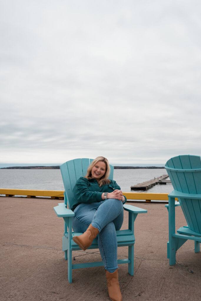 St. Louis Photographer, Hailey Lunsford, pictured on Prince Edward Island, Canada, sitting in a blue beach chair on a boardwalk in front of the ocean. Hailey has her legs crossed, wearing blue jeans, brown boots, a green sweater, and a rose gold apple watch. 