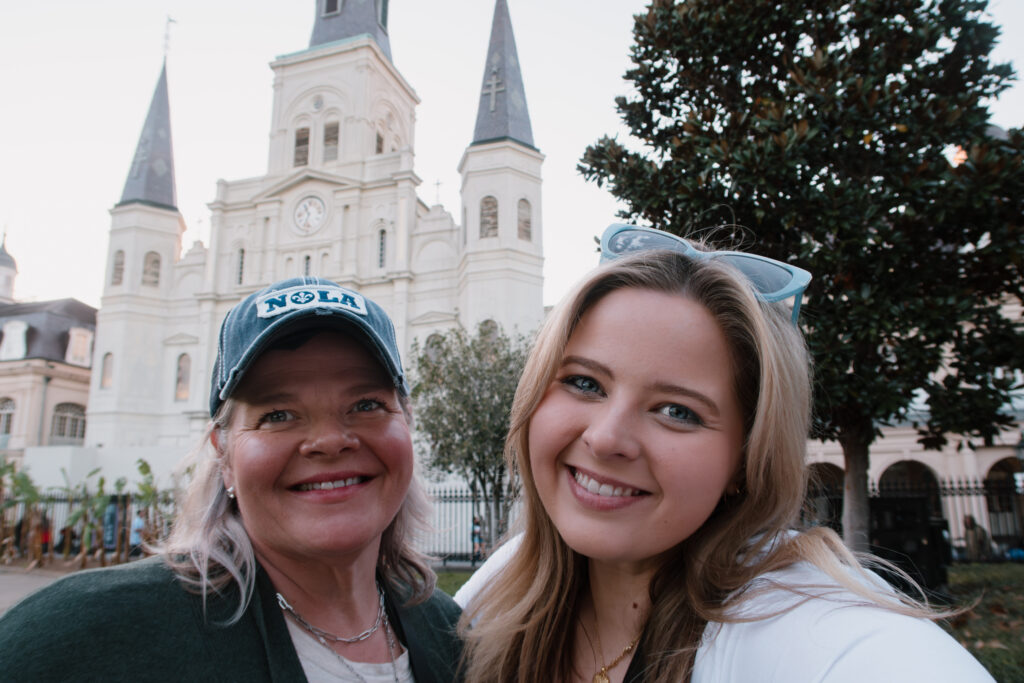 St. Louis photographer, Hailey Lunsford, pictured wearing a white longsleeve t-shirt and blue sunglasses standing in front of Jackson Square in New Orleans, LA next to her mom, who is wearing a New Orleans hat, white shirt, and green cardigan. 