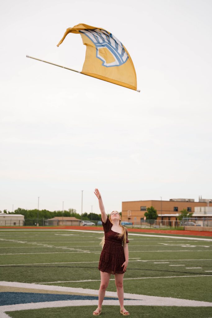 Image of senior photo session captured by St. Louis senior photographer, Hailey Lunsford. Woman stands in a red polka-dot dress throwing a yellow school flag in the air.