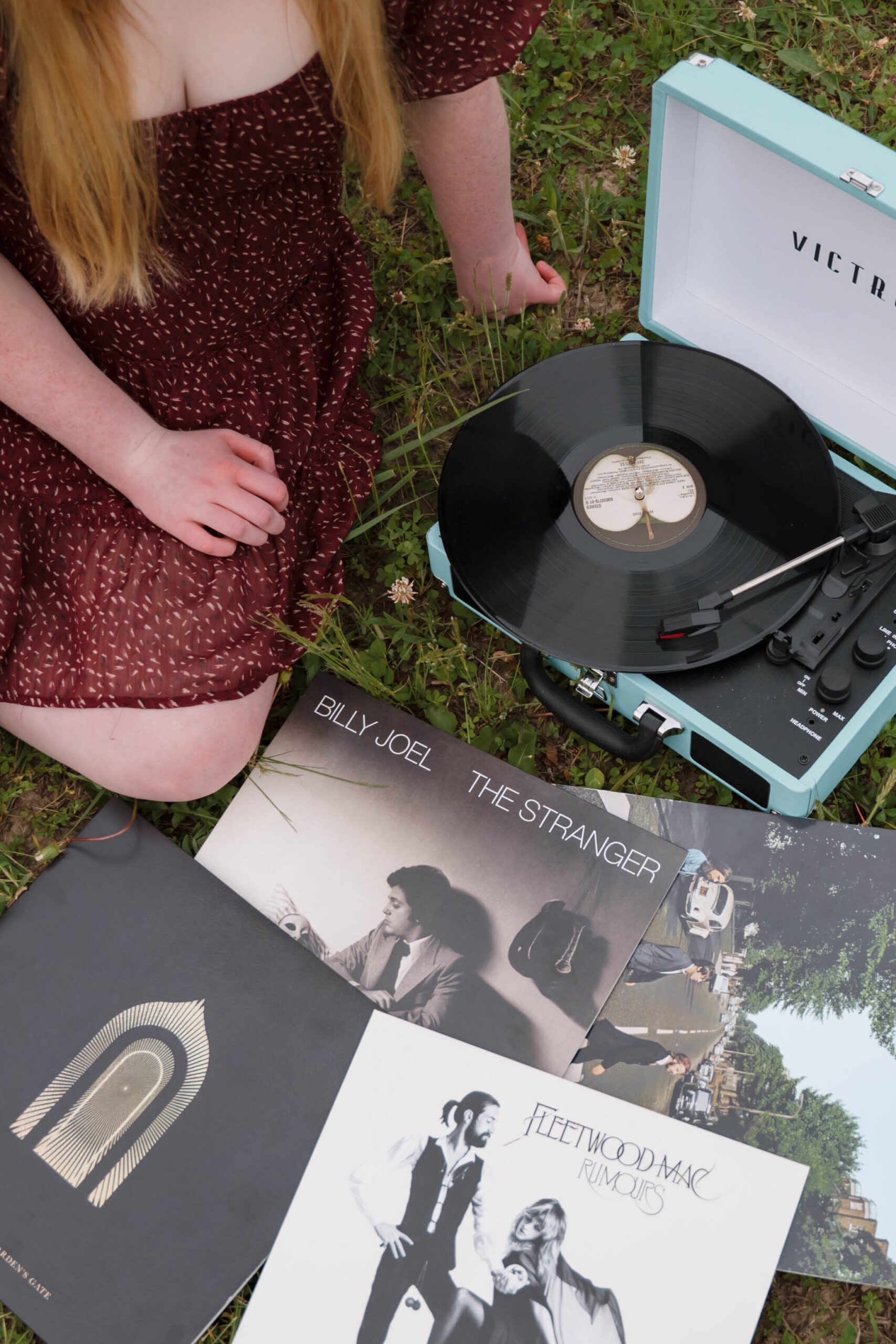 Image of senior photo session captured in St. Louis, MO by senior photographer, Hailey Lunsford. Girl with red hair sits wearing a red polka-dot dress accompanied by a blue record player and classic records such as Billy Joel, Fleetwood Mac, and The Beatles.