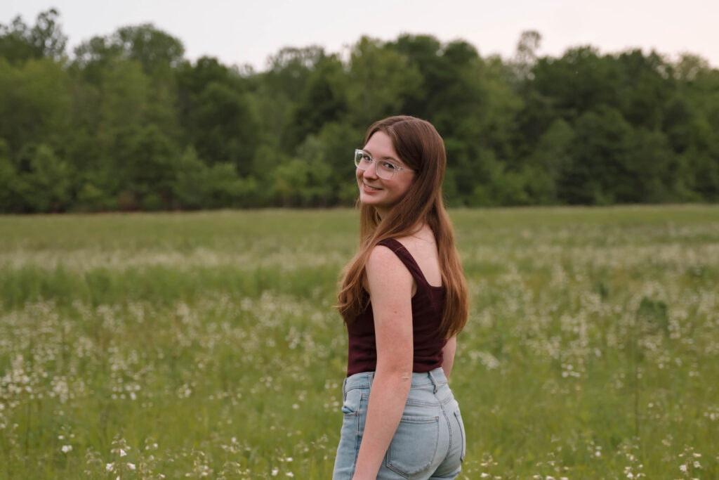 Photo of a senior during a senior photo session in St. Charles, MO captured by Hailey Lunsford Photography. Woman stands in a field with greenery in the background, looking back at the camera. She has long brown hair, clear glasses, wearing a maroon tank top and jeans. 