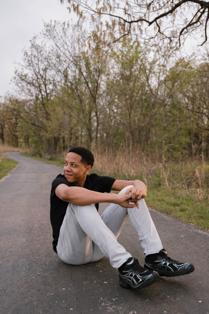 Image captured of a senior photo session by Hailey Lunsford Photography. Man sits along trail at a park in Wentzville, MO, wearing a black t-shirt, khaki pants, and black tennis shoes.