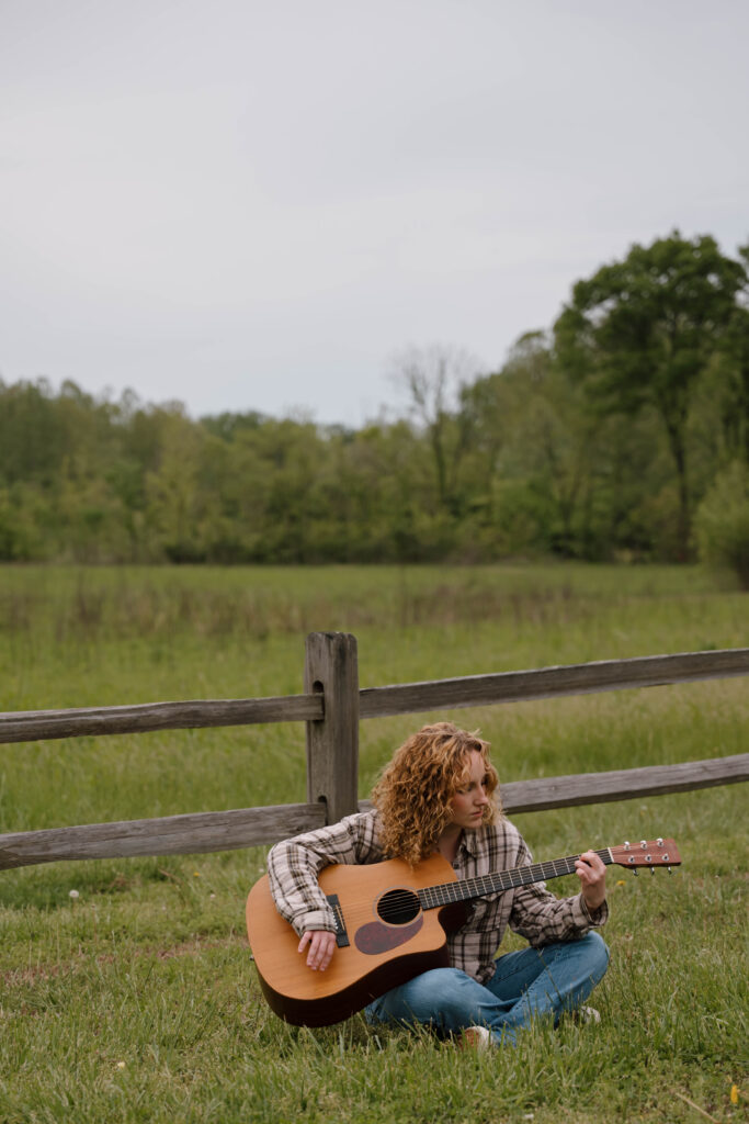 Image of a senior photo session in St. Charles County, MO, captured by senior photographer, Hailey Lunsford Photography. Woman sits in front of a wood fence with greenery in the background, strumming a brown guitar. She has strawberry blonde, curly hair, wearing a brown plaid long-sleeve shirt and jeans.