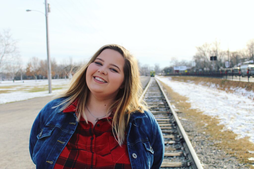 Image of Hailey Lunsford, St. Louis senior photographer, during her own senior portraits in 2018. She stands in front of the train tracks at Historic Main Street. Snow covers the grass in the background. Hailey has dirty blonde hair, wearing a red flannel and a jean jacket.
